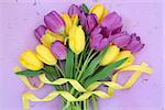Yellow and purple tulip flower bouquet with ribbon over mottled lilac background.