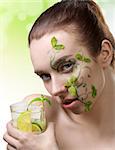 beauty portrait of cute brunette girl with creative green make-up and some mint leaves on the face, taking a cocktail mojito in hand