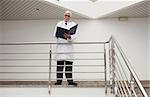 Doctor reading patient files leaning against railing in hospital corridor