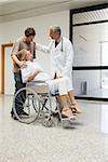 Doctor laughing with pregnant woman in wheelchair and partner in hospital corridor