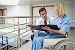 Doctor and nurse in wheelchair studying  folder in hospital corridor