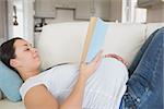 Young pregnant woman with a hand on her belly reading a book in he living room