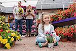 Family holding flower pots and smiling with little girl sitting on ground in garden center