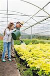 Little girl watering plants with grandfather in greenhouse