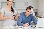 Disappointed couple in the kitchen cutting credit card and looking at finances on laptop