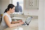 Woman using a laptop in the kitchen with burglar standing at the window