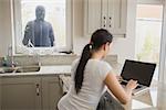 Woman on laptop in kitchen being observed by burglar through window