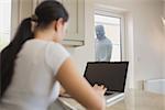 Burglar looking at woman using laptop in kitchen through glass door
