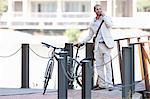 Young man by chain fence with bicycle on cell phone