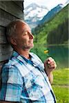 Portrait of mature man with eyes closed, holding flower, standing next to building at Lake Vilsalpsee, Tannheim Valley, Austria