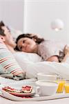 Mid adult couple lying in bed, breakfast on tray, hugging