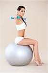 Girl lifting weights while sitting on fitness ball  isolated on baige background