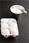 Chicken eggs in white carton with flour in metal measuring cup on dark background