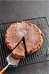 Freshly baked homemade chocolate cake on metal cooling rack with cut slice and cake server utensil on dark background