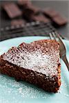 Slice of freshly baked homemade chocolate cake on plate with fork and chocolate squares in background