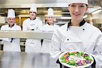 Smiling chef holding out a salad in the kitchen with team standing behind her