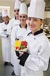Smiling chef holding fruit plate with team of Chef's in kitchen