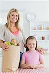 Woman and her little girl smiling at the kitchen with grocery bag on the table