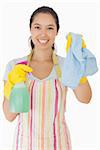 Smiling young woman holding up rag and spray bottle in apron and rubber gloves