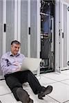 Smiling  technician working with his laptop near the server and sitting on floor of data server