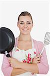 Confident young woman holding frying pan and spatula over white background