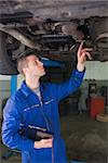 Male auto mechanic with clipboard examining under car in workshop