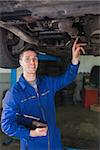 Portrait of male mechanic with clipboard examining under car in workshop