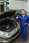 Full length portrait of mechanic by car showing thumbs up sign