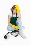Tired young maid sitting with brush and dust pan over white background