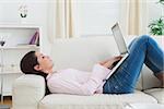 Side view of casual young woman lying on sofa and using laptop