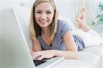 Portrait of casual young woman lying on couch and using laptop at home