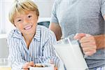 Smiling boy having cereal with father pouring milk at breakfast