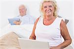 Smiling aged couple using a tablet and the laptop in the bedroom