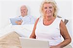 Smiling old couple using a tablet and the laptop in the bedroom