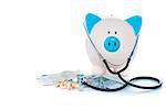 Piggy bank sitting on pile of dollars with stethoscope and pills on white background