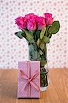 Bunch of pink roses in vase with pink gift leaning against it on wooden table