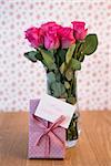 Bunch of pink roses in vase with pink gift leaning against it and mothers day card on wooden table