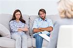 Upset couple sit on a sofa with arms crossed during a psychotherapy