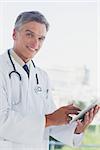 Grey haired doctor smiling and using digital tablet in a bright office