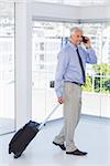 Businessman pulling suitcase and talking on phone happily in office