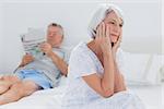 Anxious mature woman sitting on bed while husband is reading a newspaper on the background