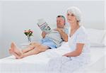 Mature woman sitting on bed while husband is reading a newspaper on the background