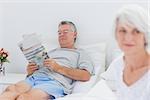 Mature man reading a newspaper in bed while his wife is sitting on bed