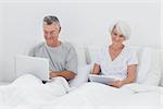 Mature man using a laptop next to wife using a tablet in bed