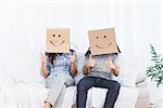 Couple sitting with cardboard boxes with smiley faces on head giving thumbs up in living room