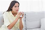 Thoughtful woman holding a cup of coffee sat on a couch