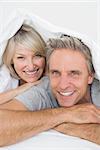 Couple smiling under the covers at the camera at home in bed