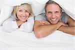 Cheerful couple smiling under the covers at the camera at home in bed