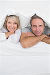 Attractive couple smiling under the covers at the camera at home in bed