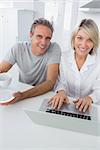 Cheerful couple using laptop in the morning looking at camera sitting at kitchen counter